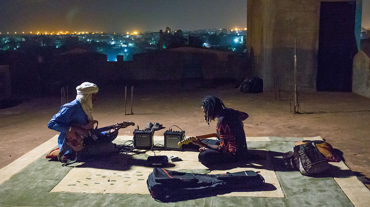 Ahmed Ag Kaedy sits on a rooftop with another guitarist at night, playing electric guitar.